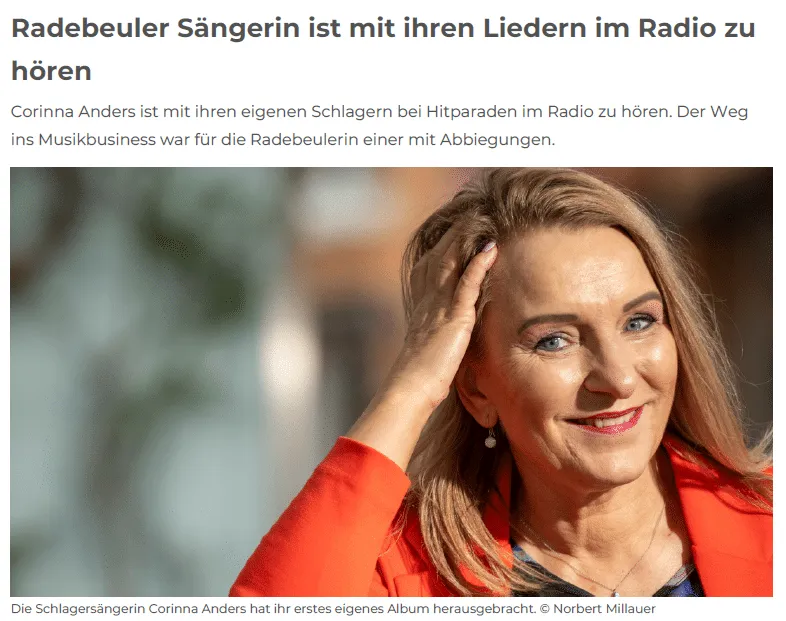 Schlagersängerin Corinna Anders im roten Blazer lächelt, strahlt ihren Kopf in die Hand und gewährt Einblicke vor unscharfer Natur im Hintergrund.